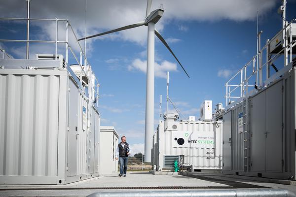 Weiße Container mit technischen Anlagen, im Hintergrund eine Windkraftanlage