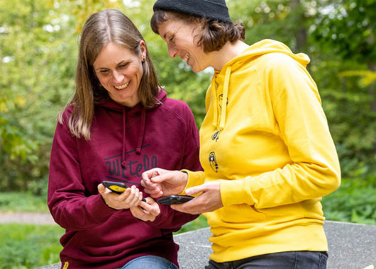 Zwei weiblich gelesen Personen unterhalten sich., eine hält ein Handy in der Hand.