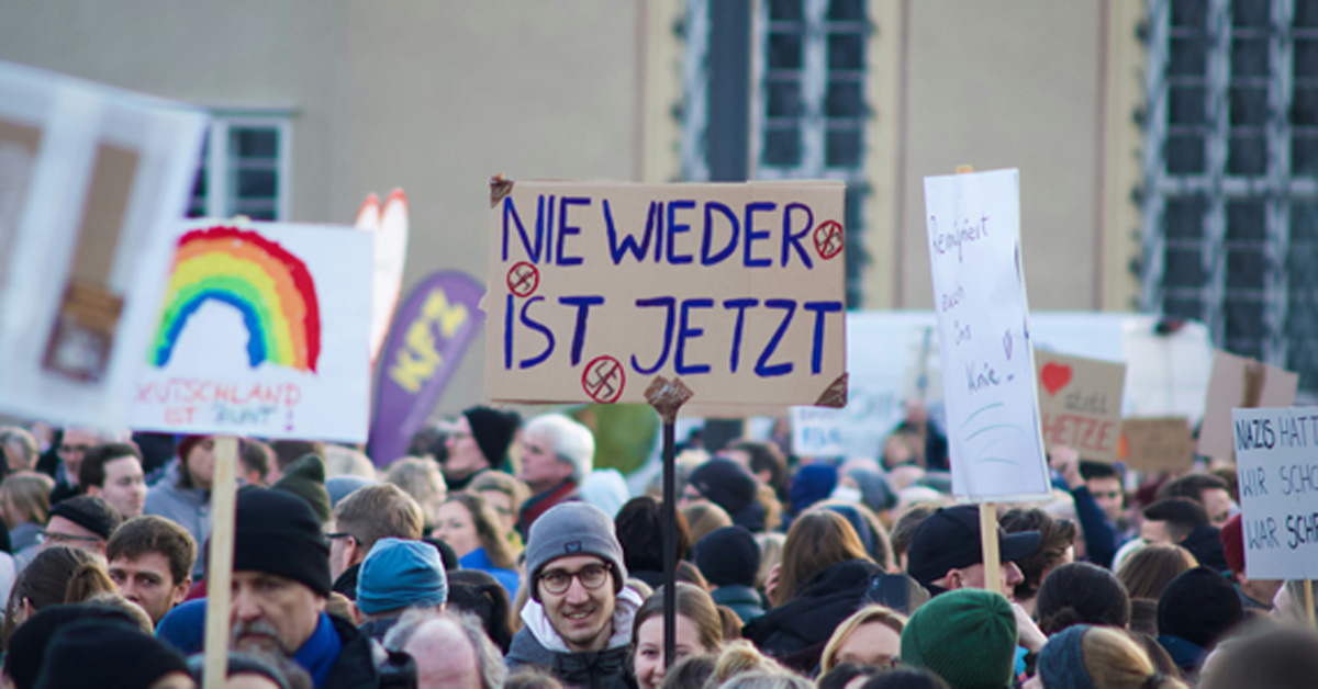 Demonstration: Ein*e Teilnehmer*in hält ein Plakat mit der Aufschrift "Nie wieder ist jetzt" und durchgestrichenen Hakenkreuzen