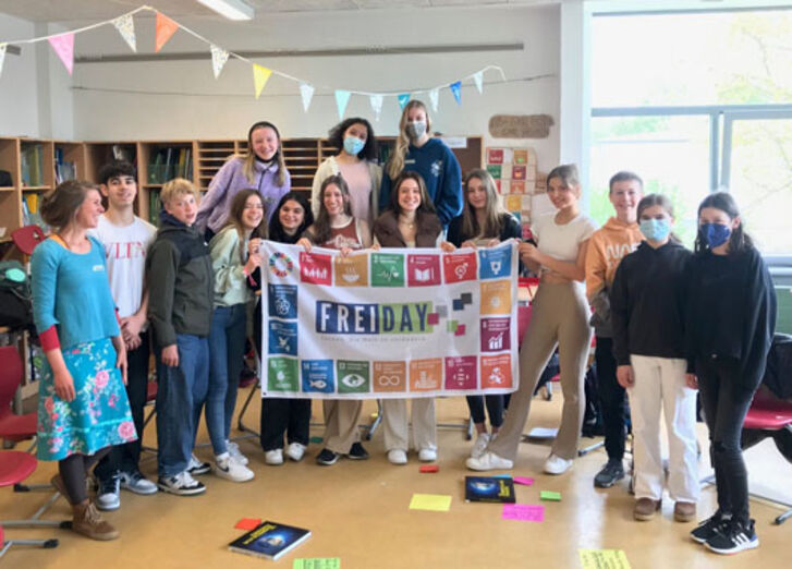 14 Schülerinnen und Schüler stehen mit ihrer Lehrerin vor einem Plakat von Schule im Aufbruch