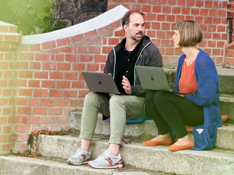 Zwei Menschen mit Laptops sitzen auf einer Eingangstreppe.