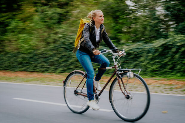 Frau fährt mit einem Fahrrad über eine Straße. Im Hintergrund Bäume und Büsche.