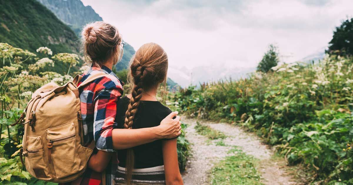 Zwei weiblich gelesene Personen, eine mit Rucksack, schauen auf ein Bergpanorama