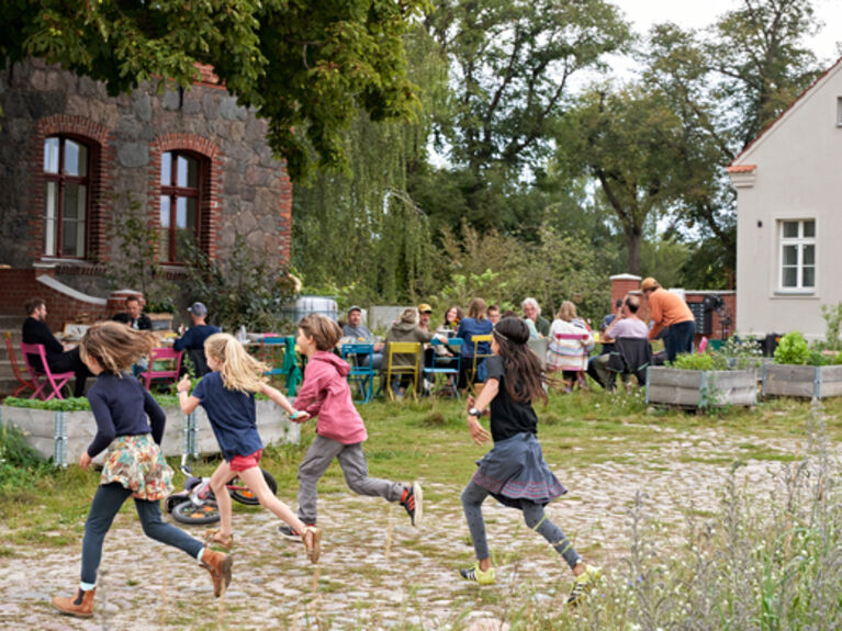 Spielende Kinder auf einer blühenden Wiese, mit älteren Häusern im Hintergrund