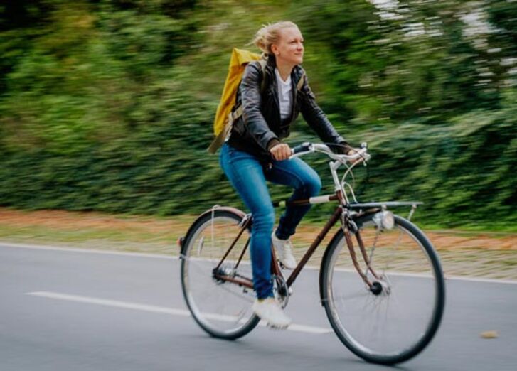 Weiblich gelesene Person fährt mit einem Fahrrad auf einer Straße vor begrüntem Hintergrund