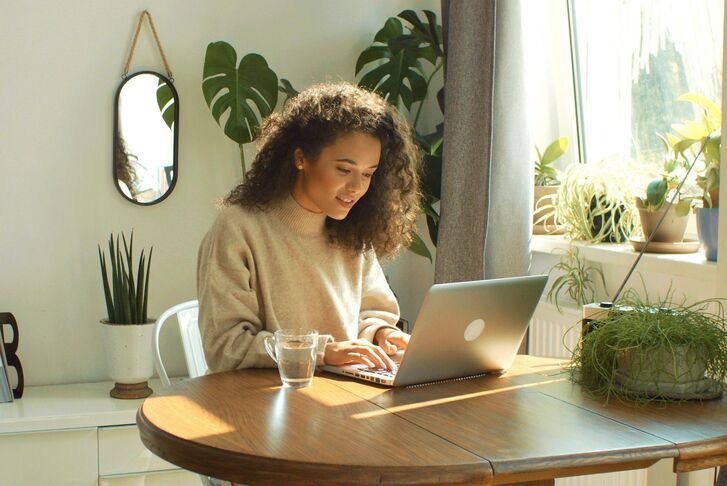 Frau mit Laptop an einem Holztisch. Im Hintergrund grüne Pflanzen.