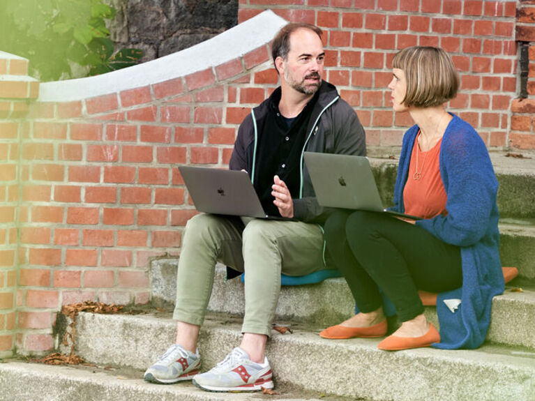 Eine Frau und ein Mann sitzen auf einer Außentreppe mit Laptops.