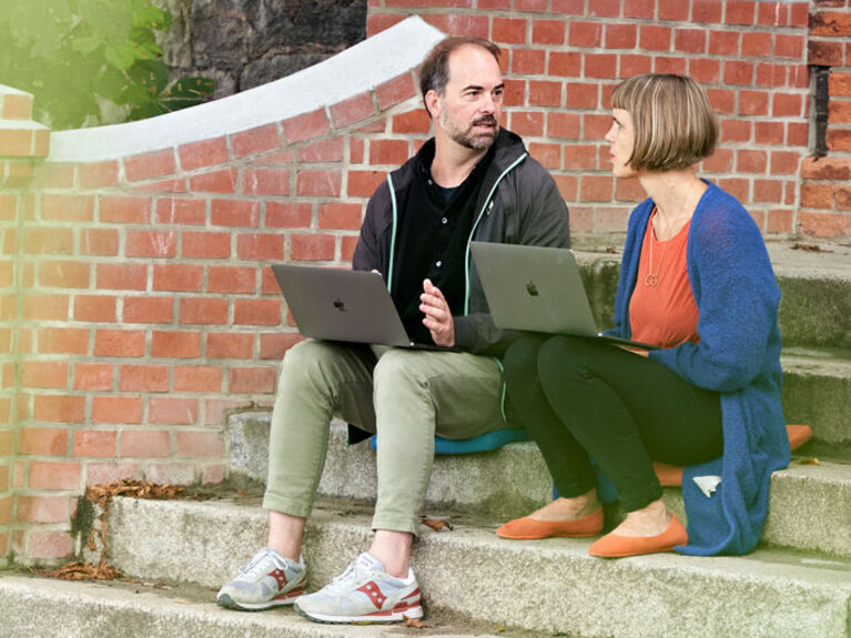 Eine Frau und ein Mann sitzen auf einer Außentreppe mit Laptops.