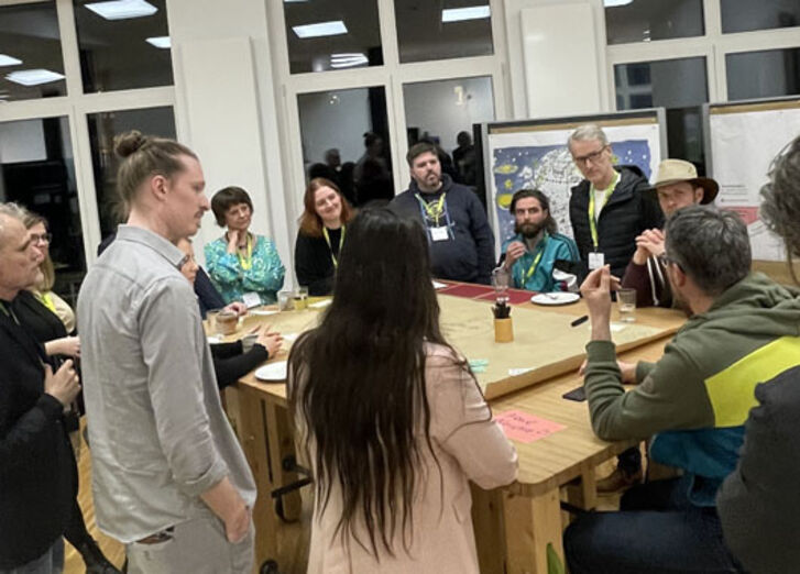 13 Personen stehen um einen Tisch, in die Gruppendiskussion vertieft