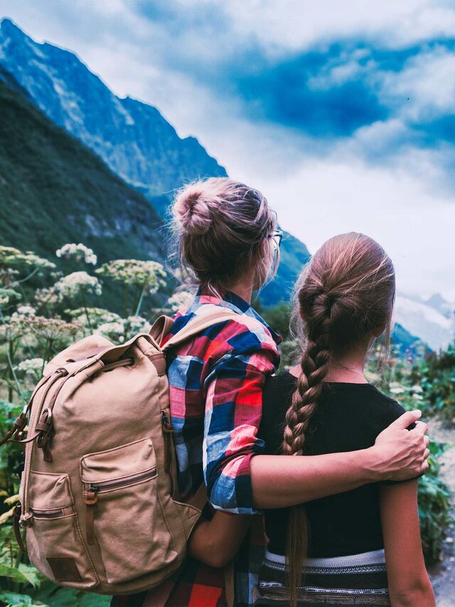 Zwei weiblich gelesene Personen, eine mit Rucksack, schauen auf ein Bergpanorama